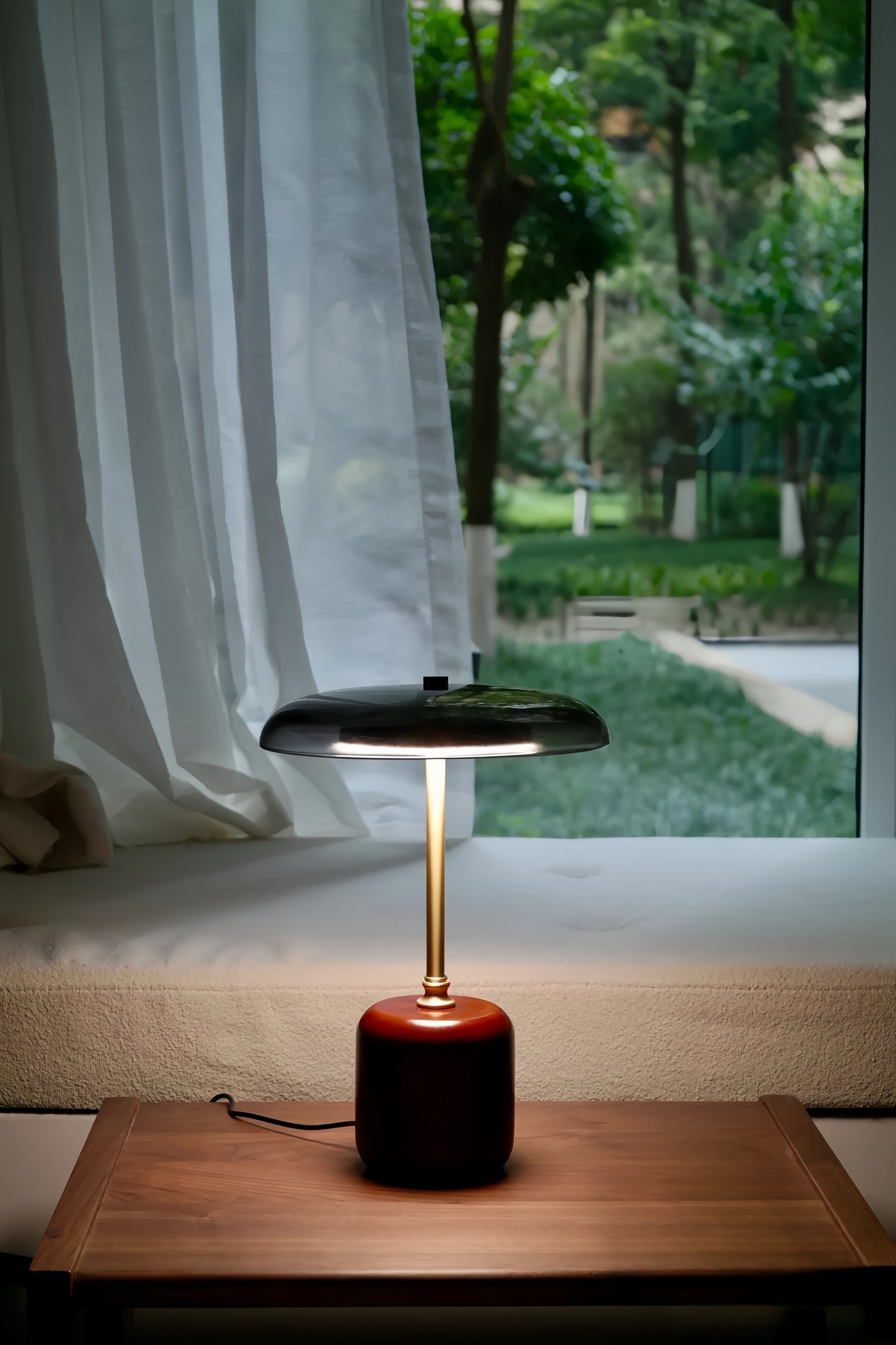American MCM table lamp with a red base on a wooden surface near a window with white curtains and a garden view.