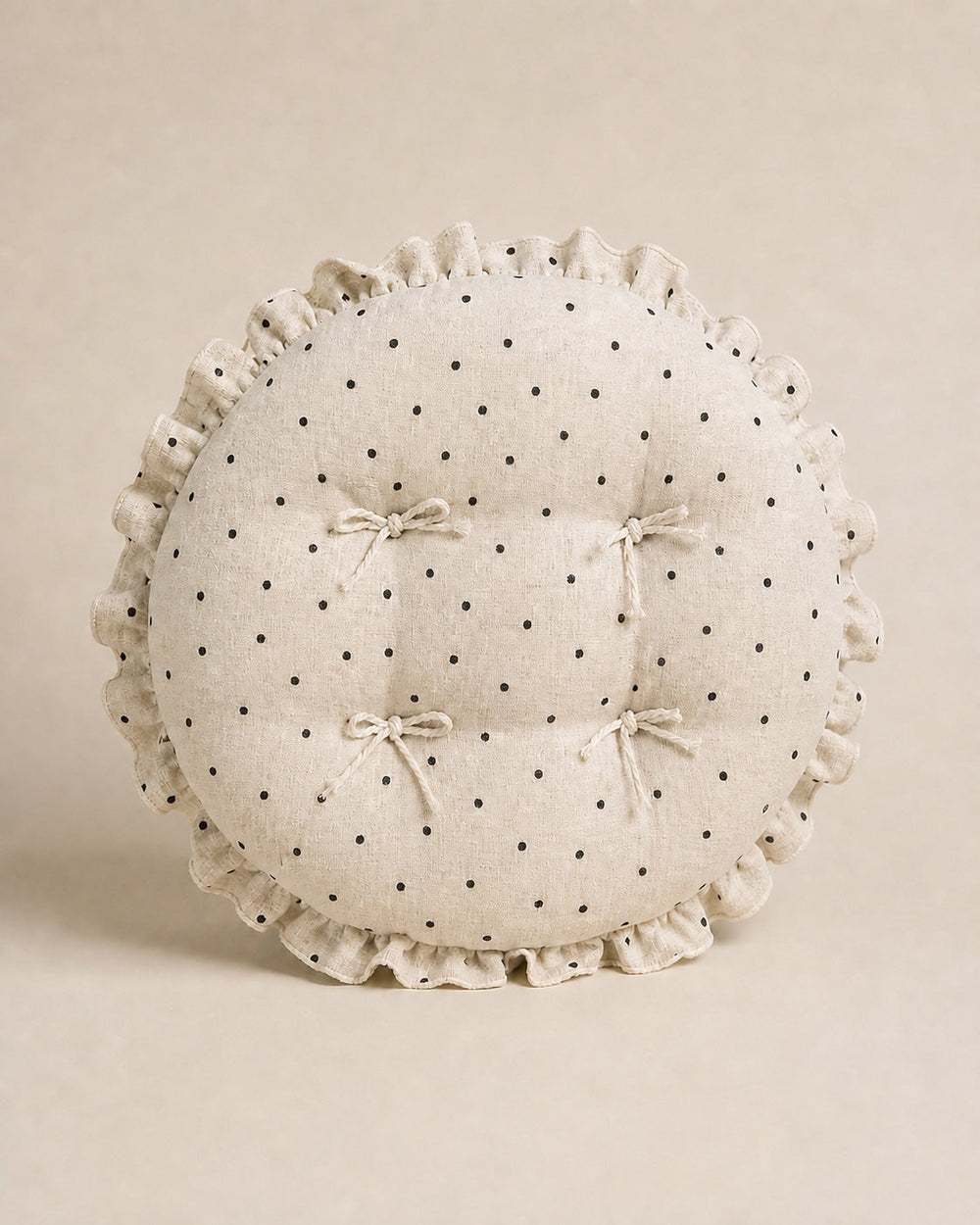 Cream polka dot round chair cushion with bow ties and a ruffled edge on a warm beige studio background.