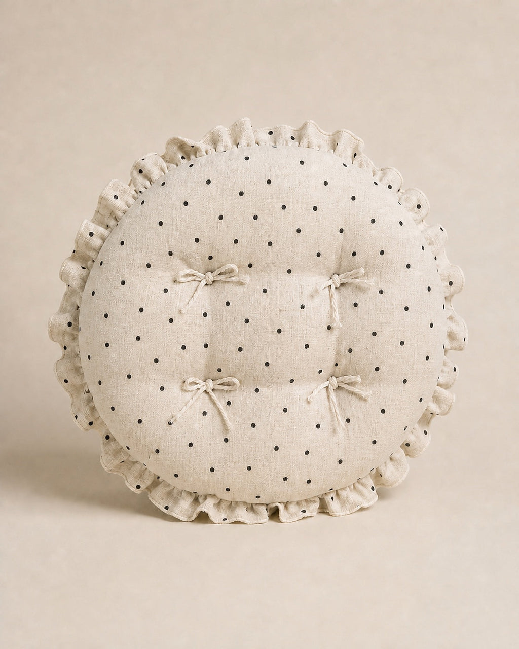 Cream polka dot round chair cushion with bow ties and a ruffled edge on a warm beige studio background.