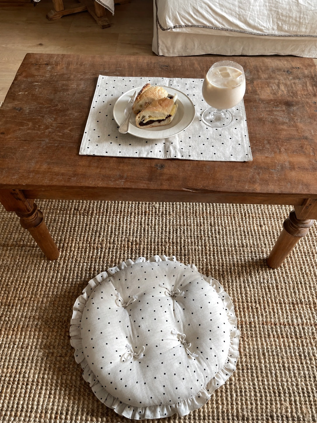 Cream polka dot round chair cushion with bow ties and a ruffled edge beside a rustic coffee table set with a drink and pastry.