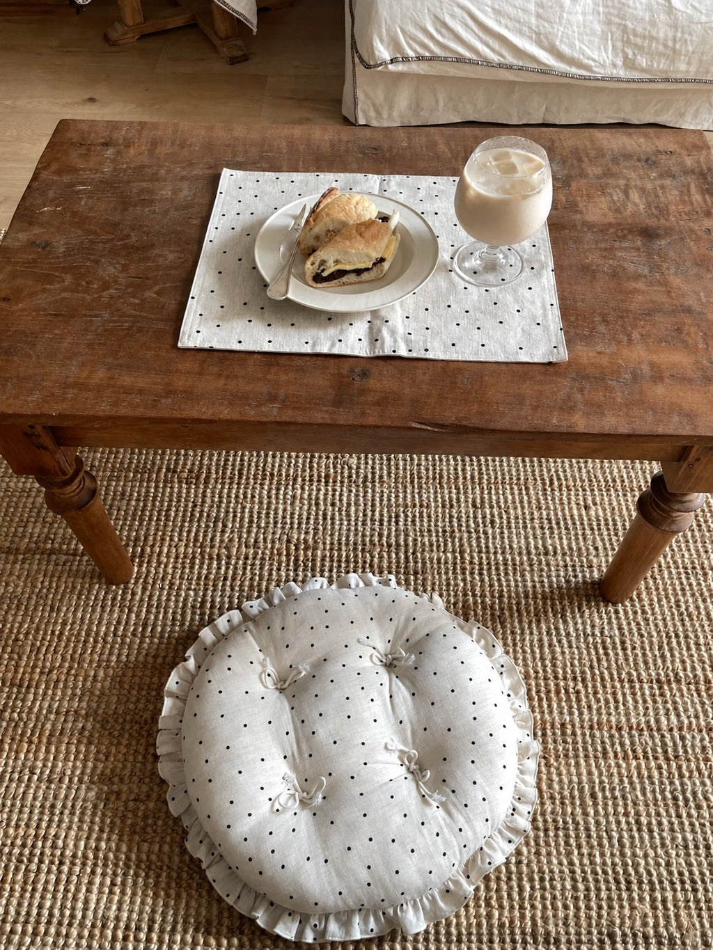 Cream polka dot round chair cushion with bow ties and a ruffled edge beside a rustic coffee table set with a drink and pastry.