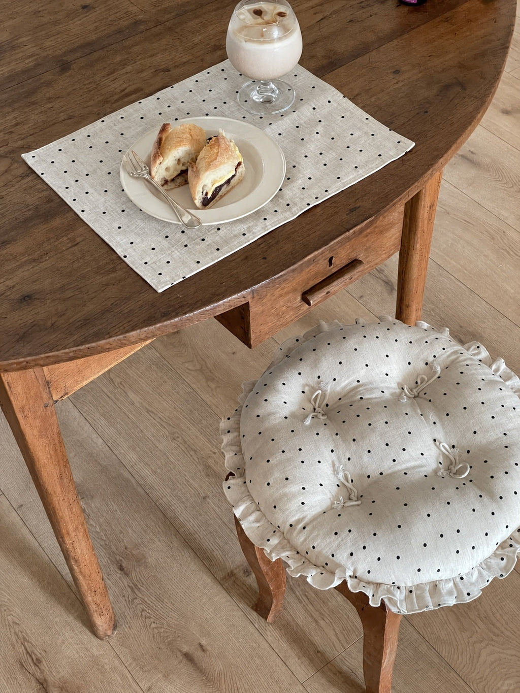 Cream polka dot round chair cushion with bow ties and a ruffled edge on a wooden chair beside a rustic dining table.