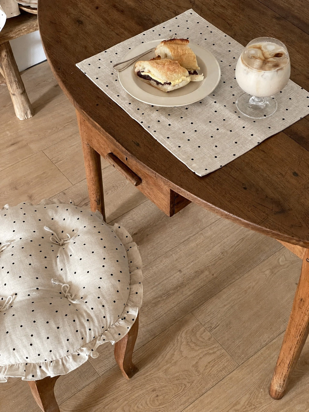 Cream polka dot round chair cushion with bow ties and a ruffled edge styled on a wooden dining chair beside a table.