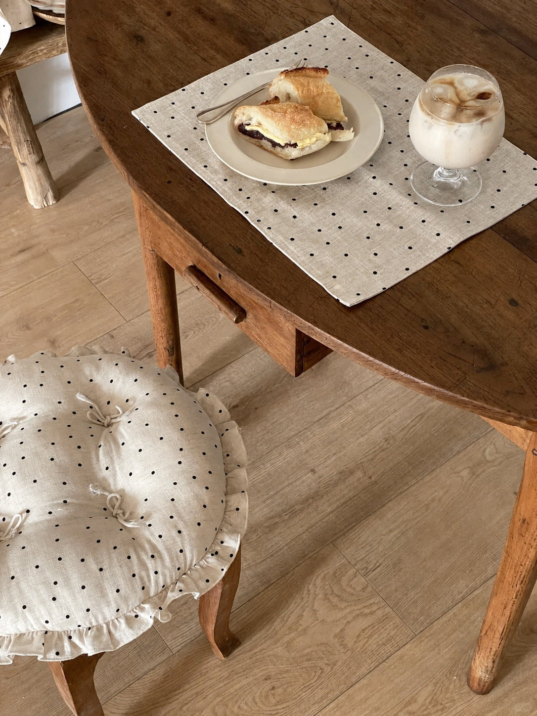 Cream polka dot round chair cushion with bow ties and a ruffled edge styled on a wooden dining chair beside a table.