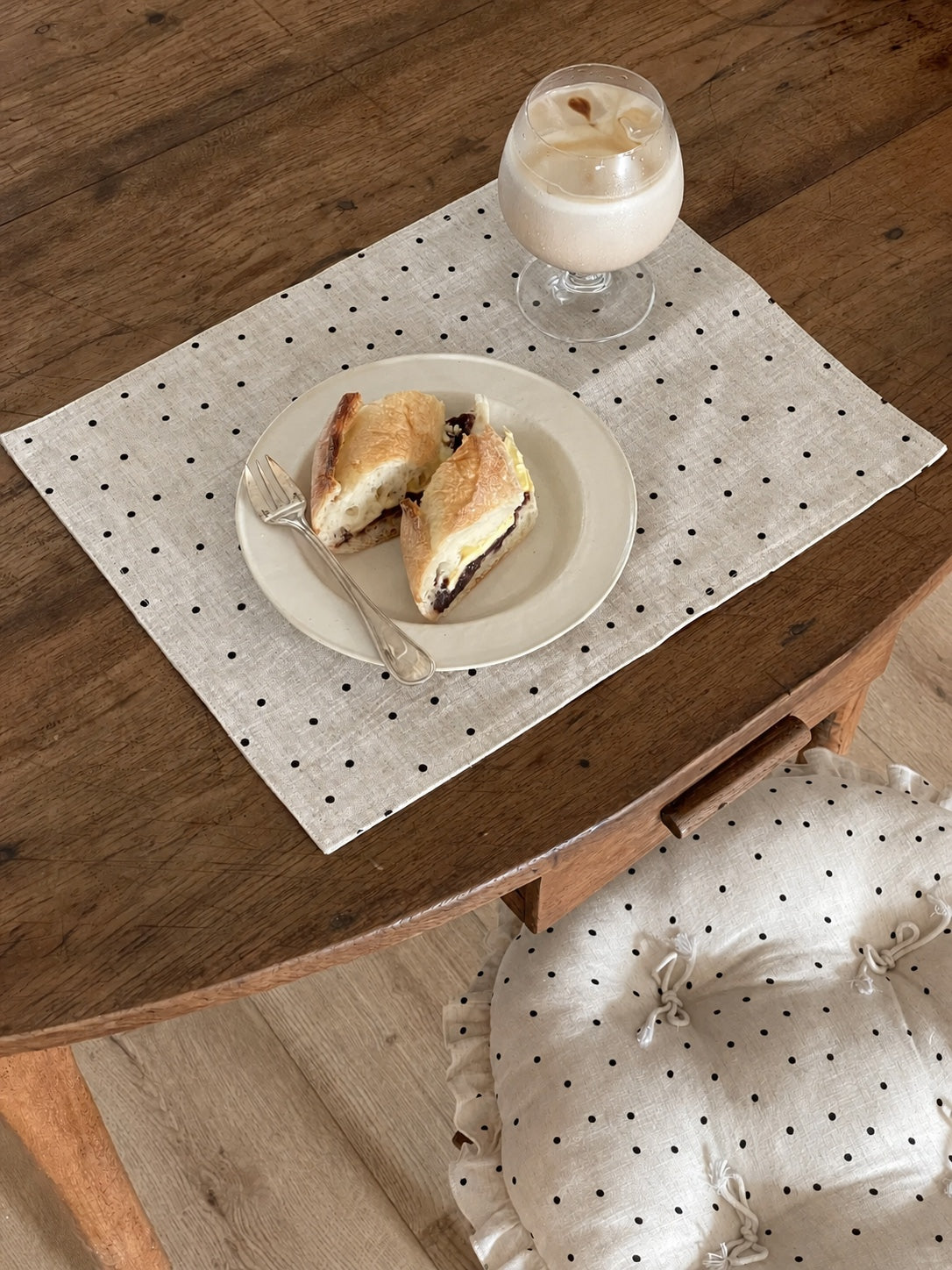 Cream polka dot round chair cushion with bow ties beside a rustic dining table with a polka dot placemat, pastry, and iced drink.