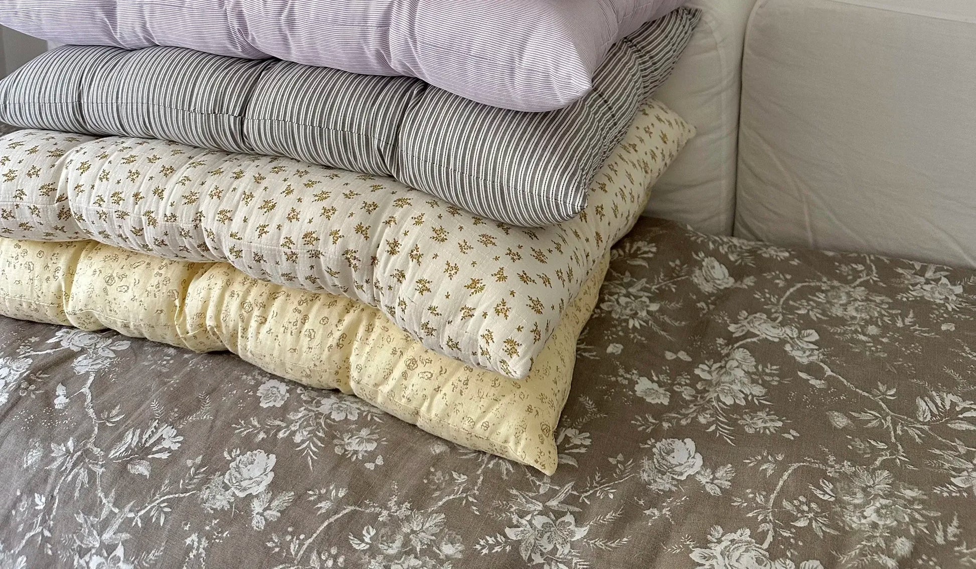 Mixed French vintage patterned pillow cushions stacked on floral bedding in a cozy room.