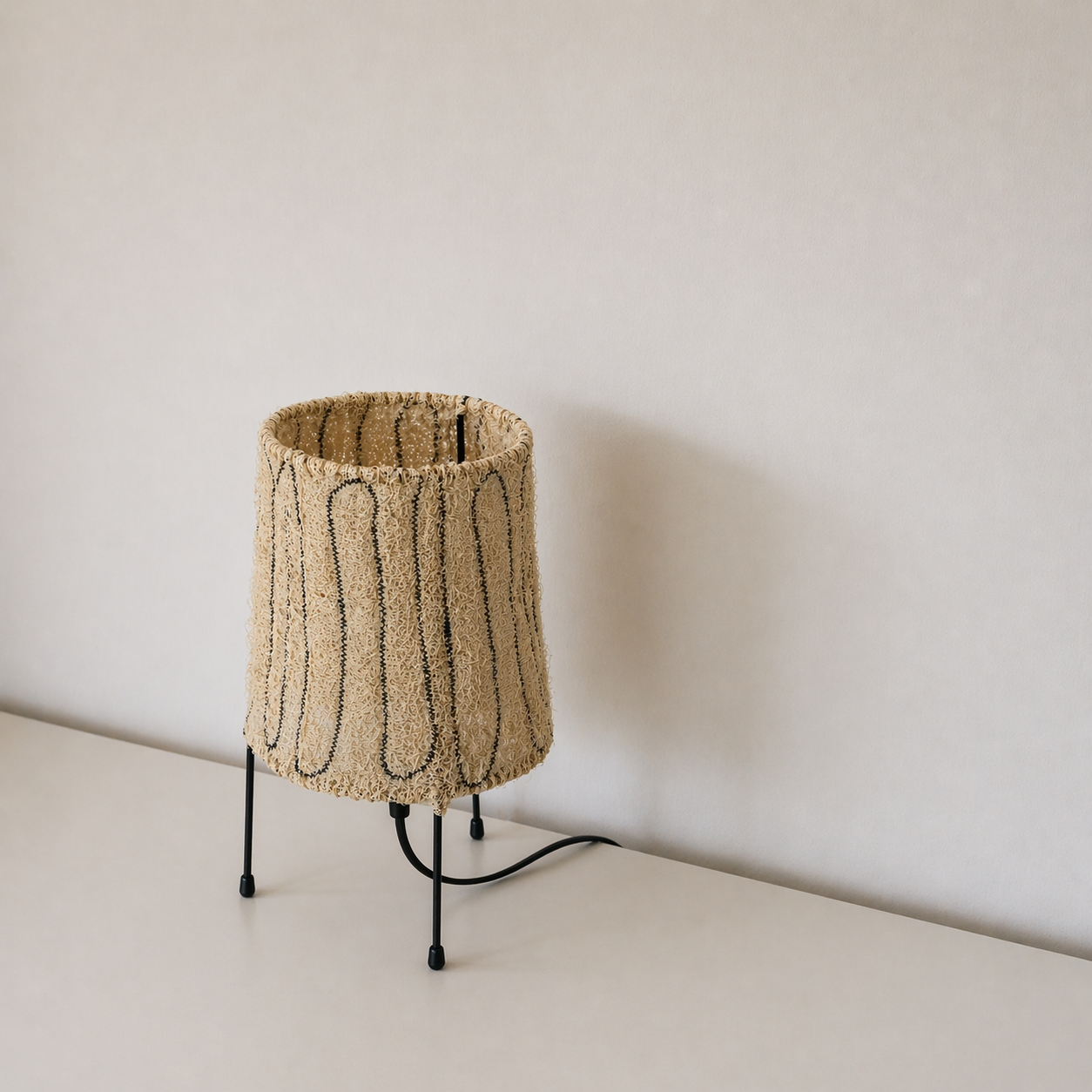 Loop loofah table lamp styled on a minimal tabletop, featuring a handmade natural fiber shade and black tripod legs.