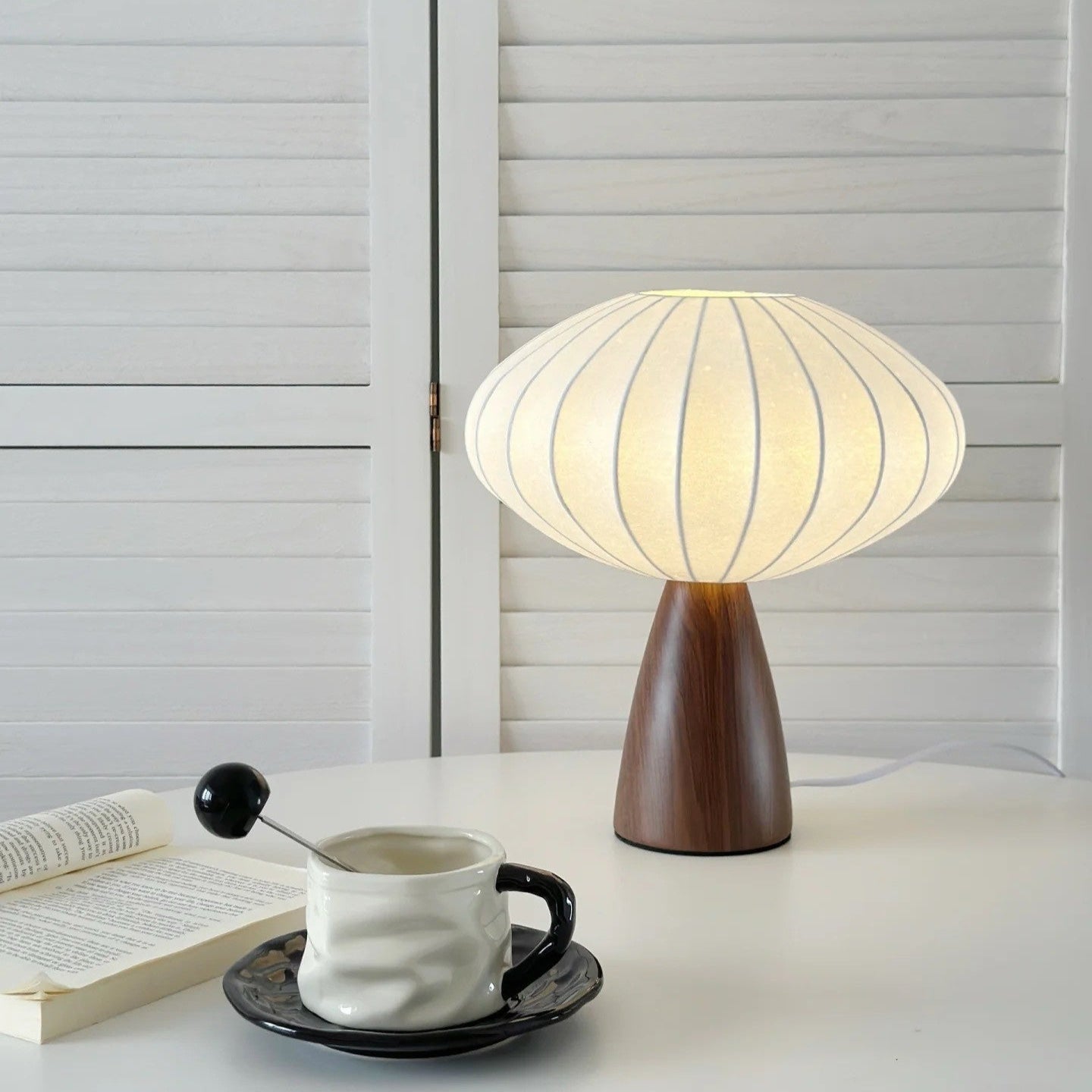 Mid-century modern table lamp with pleated fabric shade and walnut base, on a white desk with ceramic coffee cup and open book