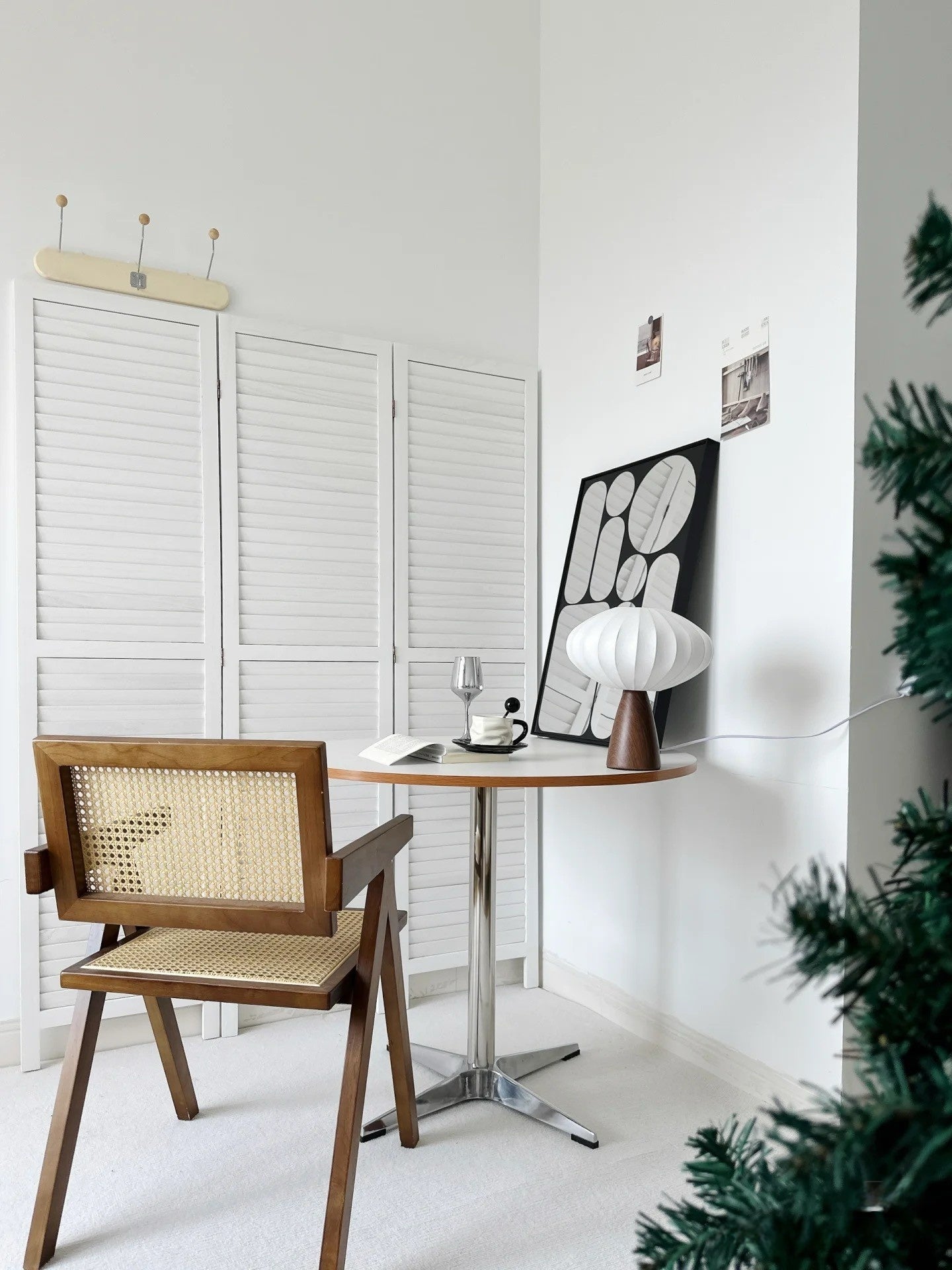 Minimalist mid-century modern reading nook with wooden cane chair, round table, pleated fabric table lamp, and decorative mirror against white shutter doors